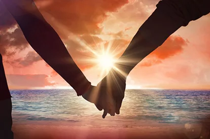 A couple is holding hands looking at the ocean and a beautiful orange sunset.